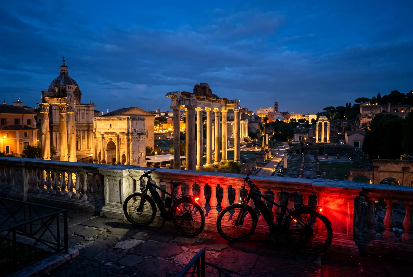 Floodlit Roman Forum at dusk with two parked e-bikes at the Capitoline balustrade