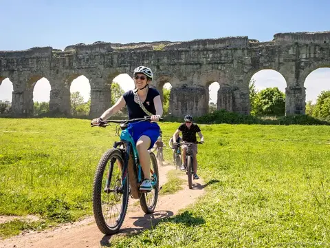 Full-suspension e-bike on a gravel lane in the Appia Antica park with aqueduct arches behind